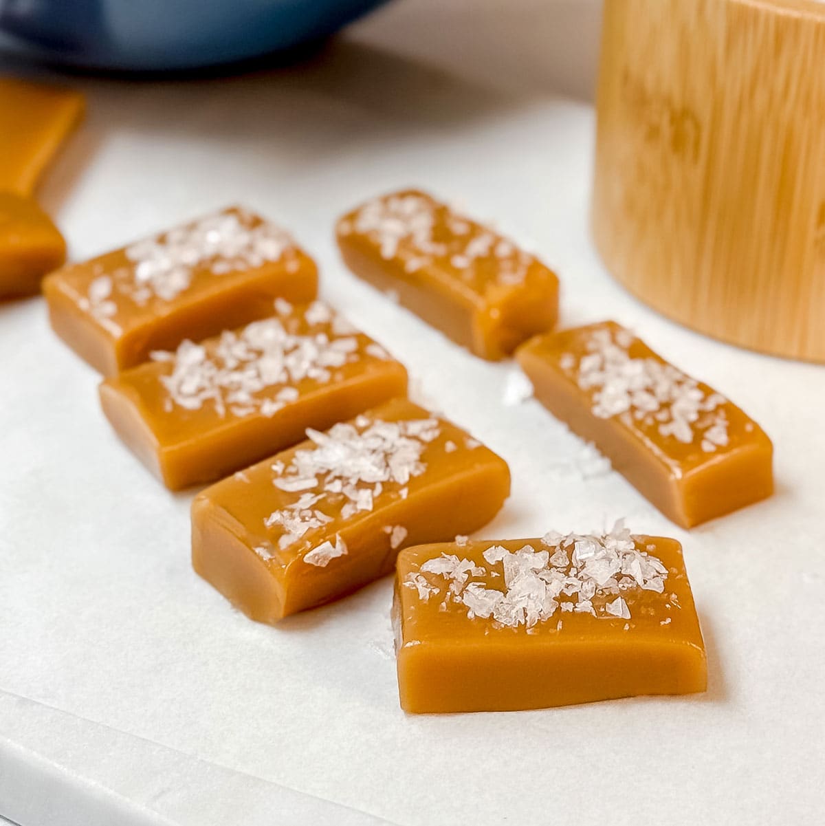 Square image of caramel candy squares topped with flaky sea salt on a parchment lined surface with wrapped candies is the background.