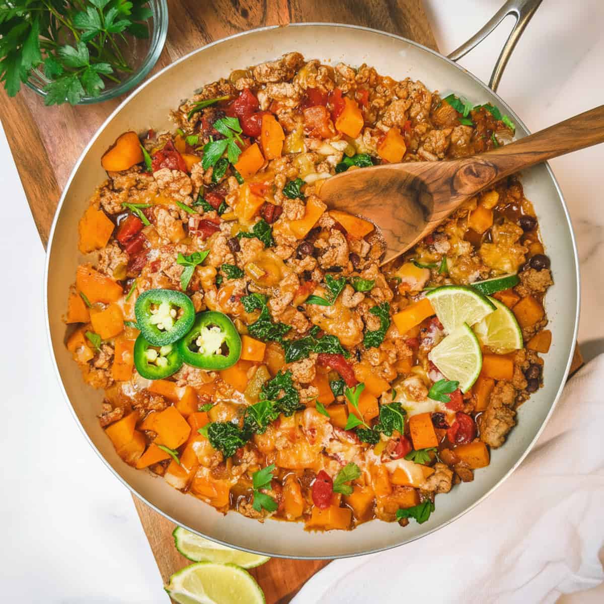 Square image of Southwestern ground turkey butternut skillet on a wooden board with a spoon and lime and jalape&ntilde;o slices to garnish.