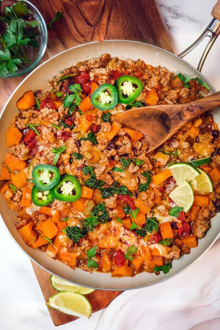 Southwestern ground turkey butternut skillet on a wooden board with a spoon and lime and jalapeño slices to garnish.