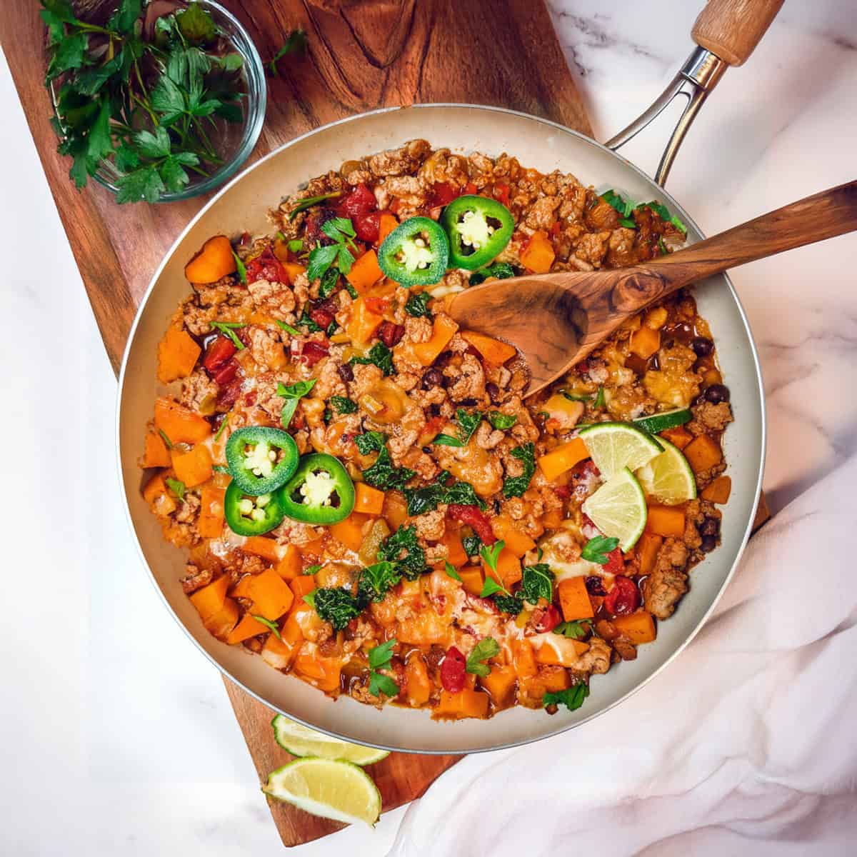 Southwestern ground turkey butternut skillet on a wooden board with a spoon and lime and jalapeño slices to garnish.