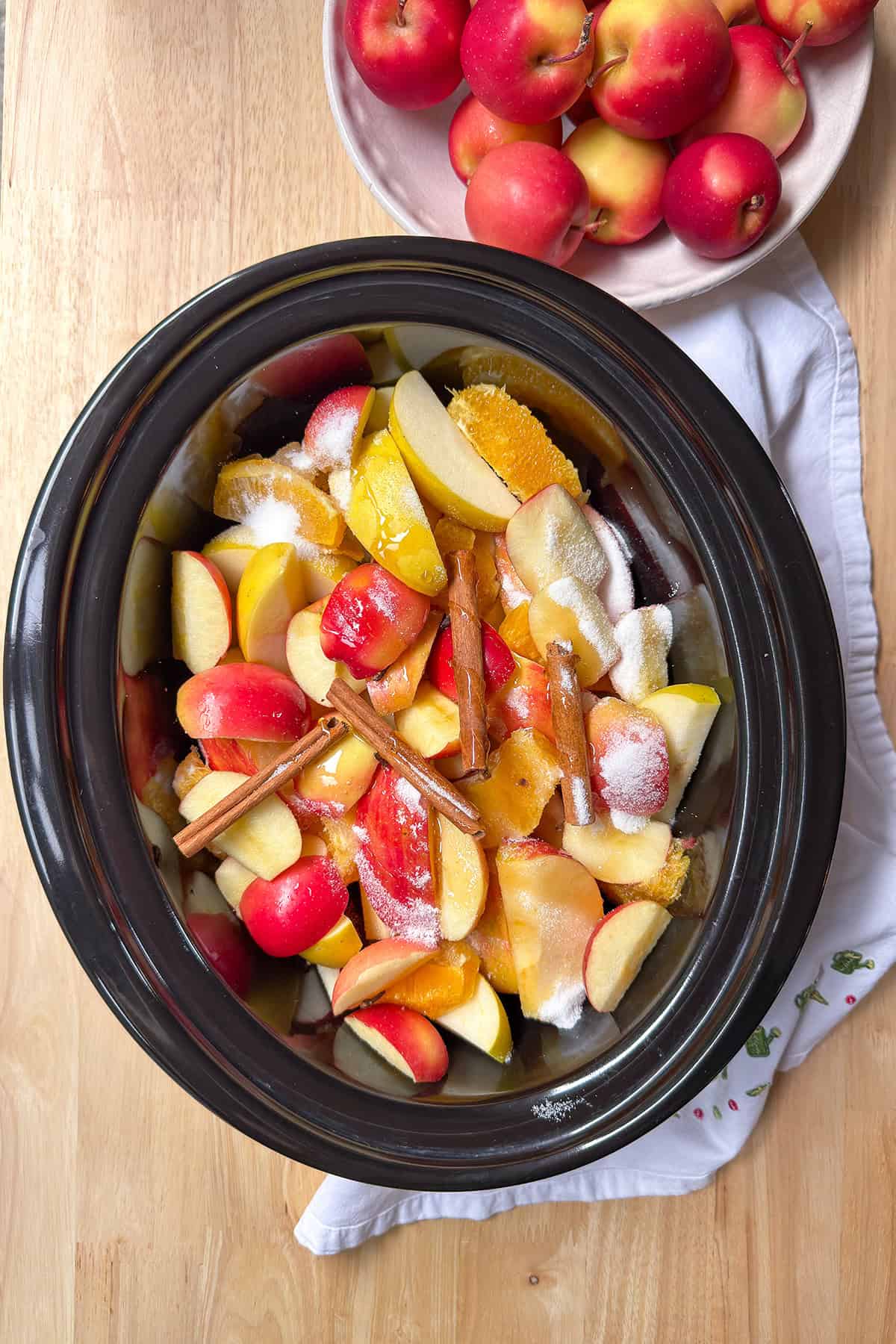 Sliced apples, oranges, sugar, seasonings, and cinnamon sticks in a slow cooker insert.
