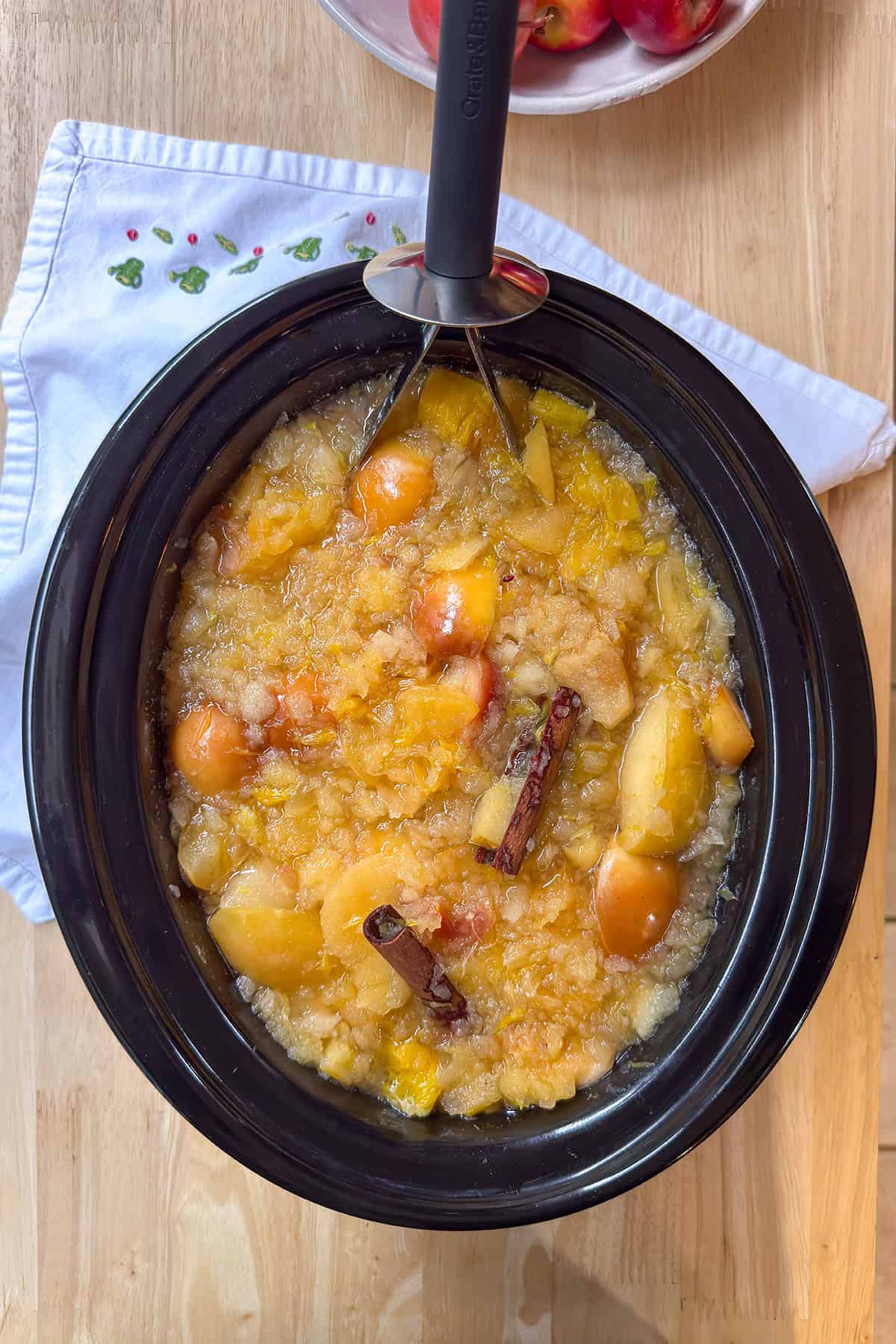 Mashed apples and oranges in a slow cooker insert with a potato masher.