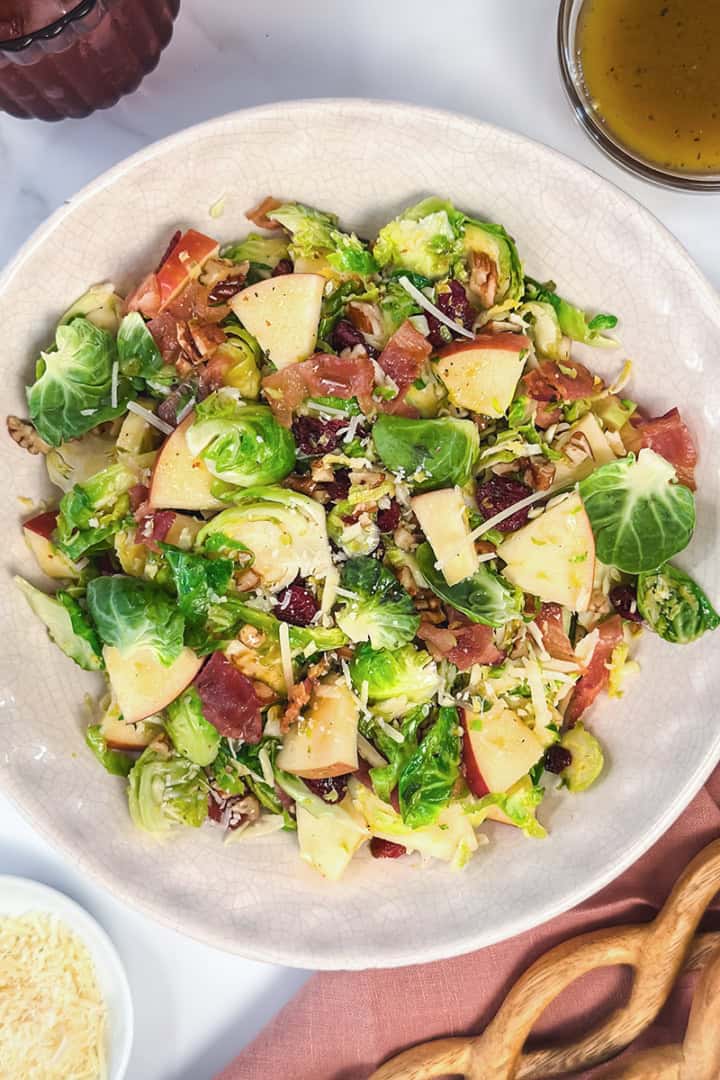 Horizontal image featuring Warm Brussels Sprouts Salad with a light serving linen, vinaigrette, and Parmesan cheese on the side.