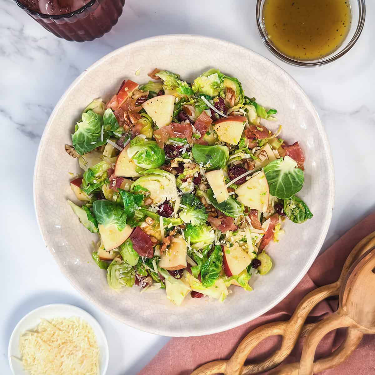 Horizontal image featuring Warm Brussels Sprouts Salad with a light serving linen, vinaigrette, and Parmesan cheese on the side.