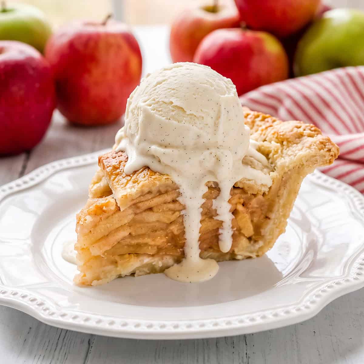 Square image of sliced apple pie with fresh apples in the background and a sliced apple pie.