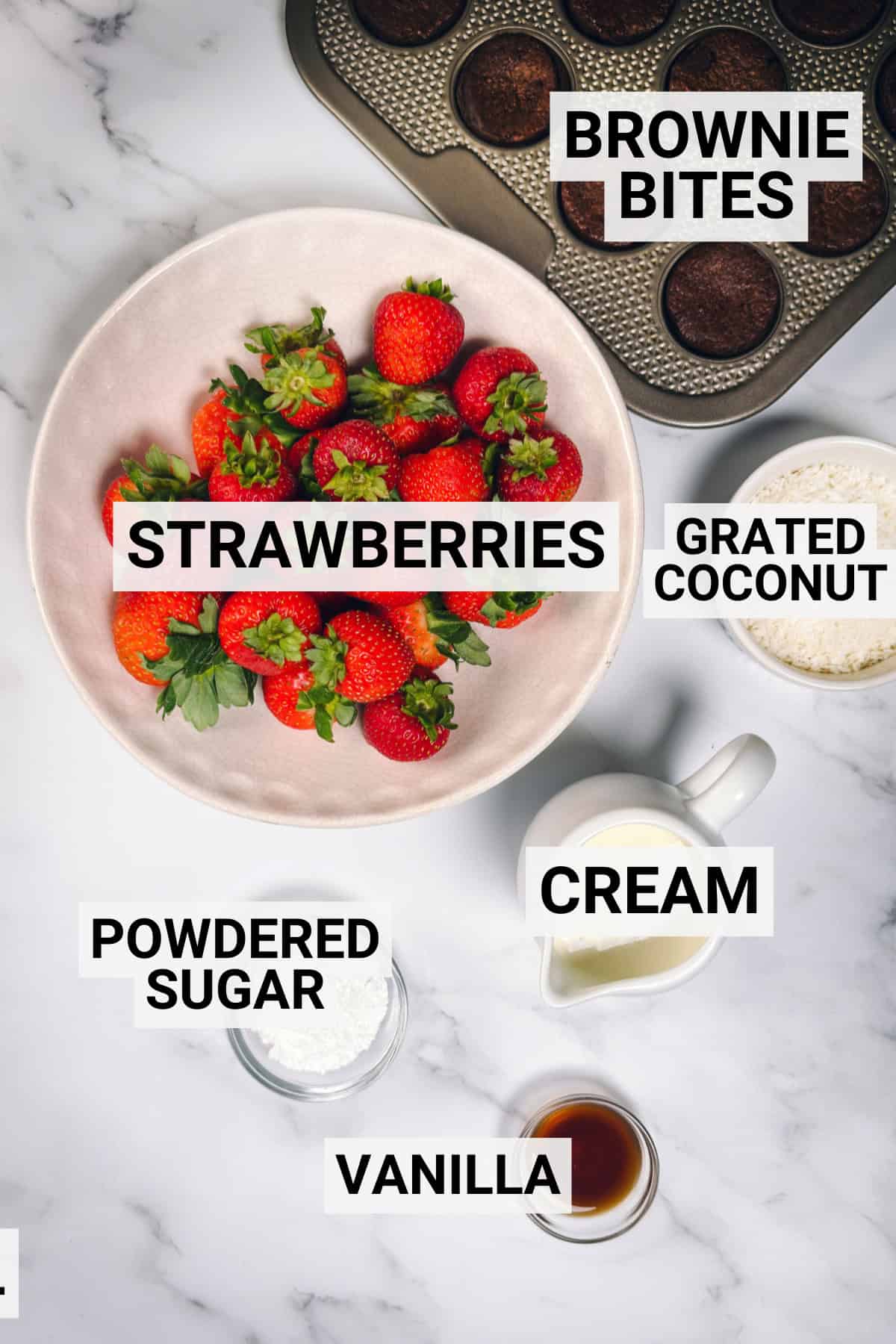 Ingredients for Santa Hat Brownie Bites including strawberries, brownie bites, whipped cream, powdered sugar, vanilla, and grated coconut arranged on a marble surface.
