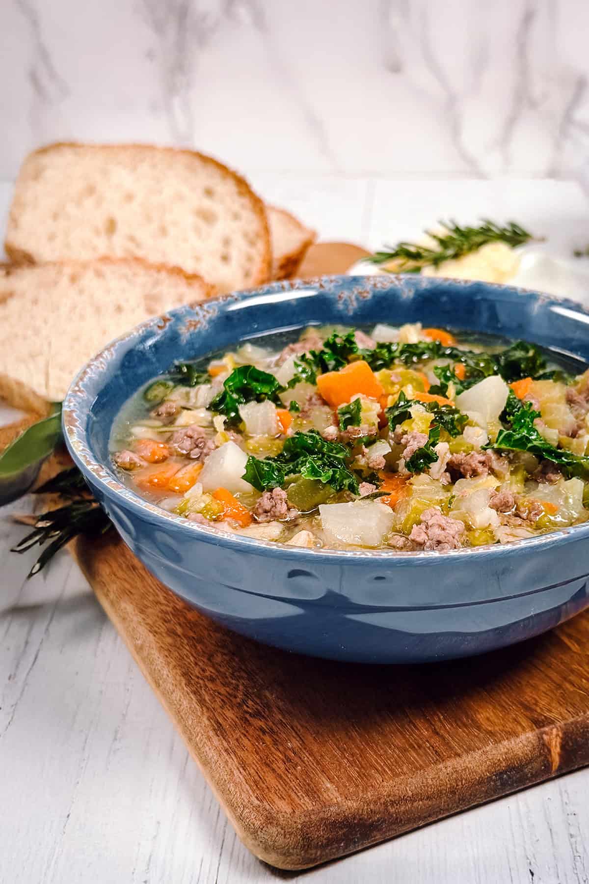 Closeup view of lamb and barley soup in a bowl on a wooden serving tray with fresh rosemary in the background.