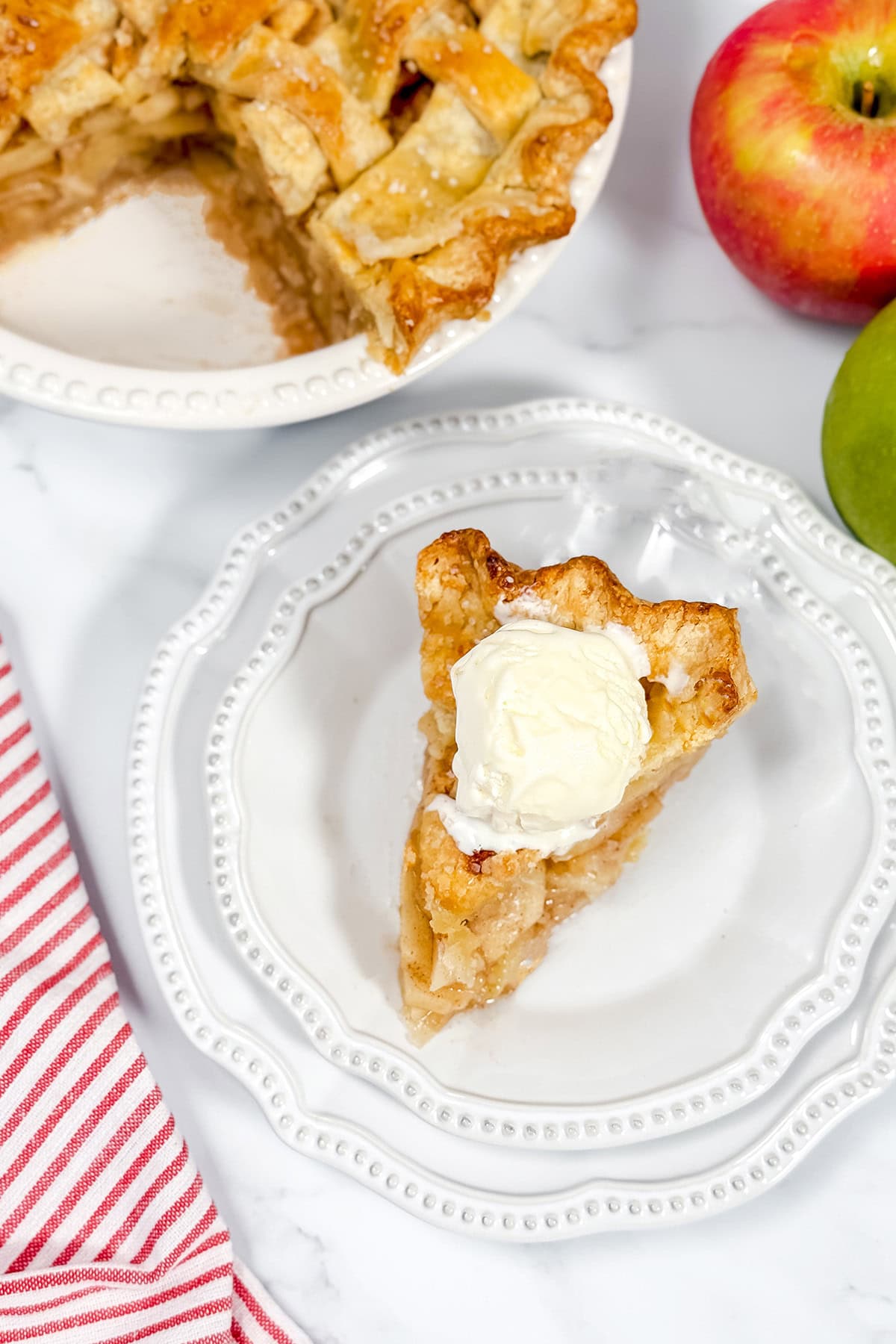 Top view of a slice of apple pie with vanilla ice cream on a white plate with the sliced pie in the background.