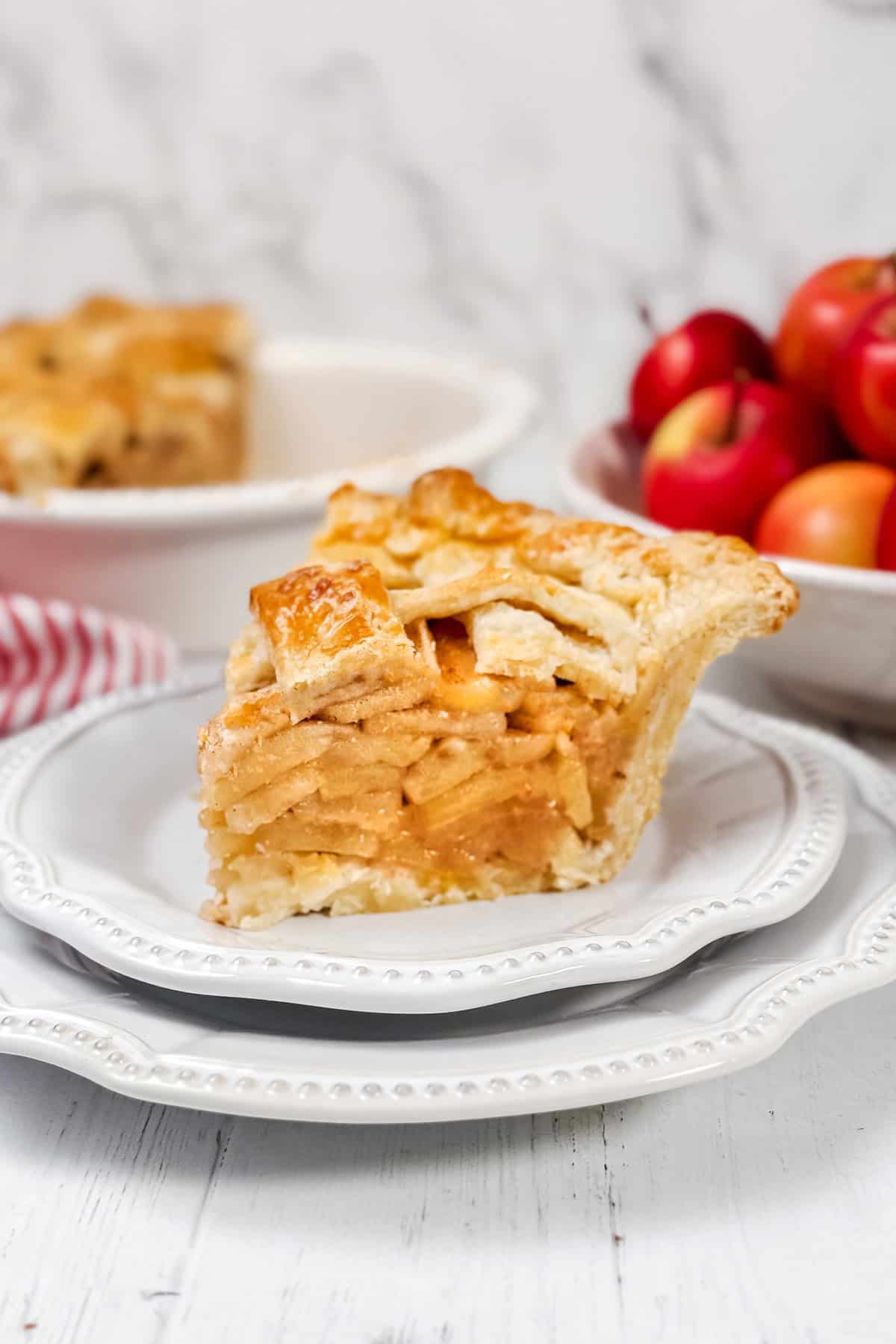 Tall slice of freshly baked apple pie on white plate with sliced pie and apples in the background.