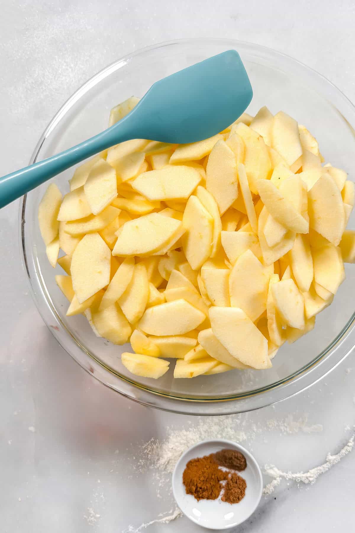 Sliced apples in a large glass bowl with sugar, flour, and spices nearby for an apple pie.