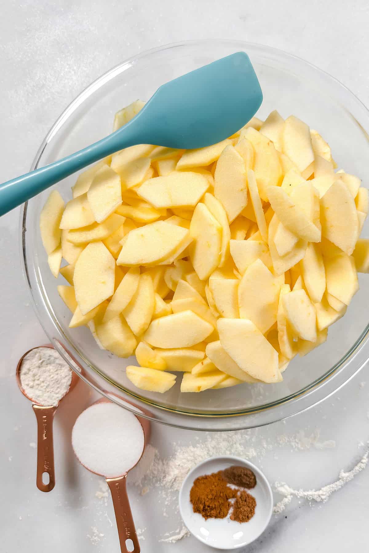 Sliced apples in a large glass bowl with sugar, flour, and spices nearby for an apple pie.