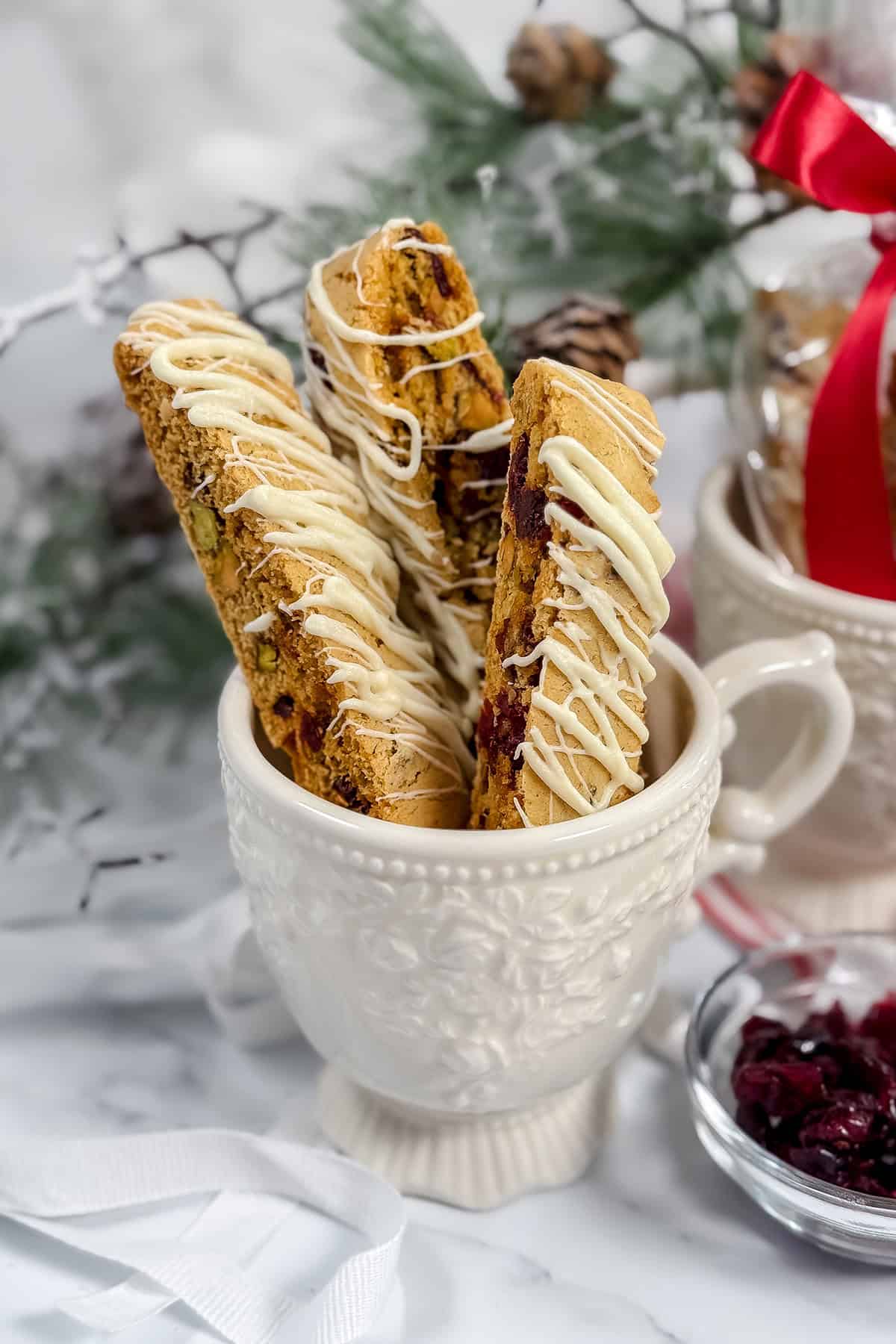 Three Cranberry Pistachio Biscotti drizzled with white chocolate are placed upright in a decorative white mug; festive decorations and dried cranberries are in the background.