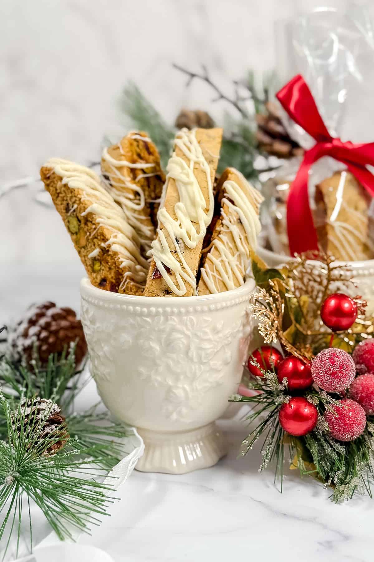 A white decorative cup holds Cranberry Pistachio Biscotti drizzled with white icing, surrounded by festive holiday greenery, pinecones, and red berries.