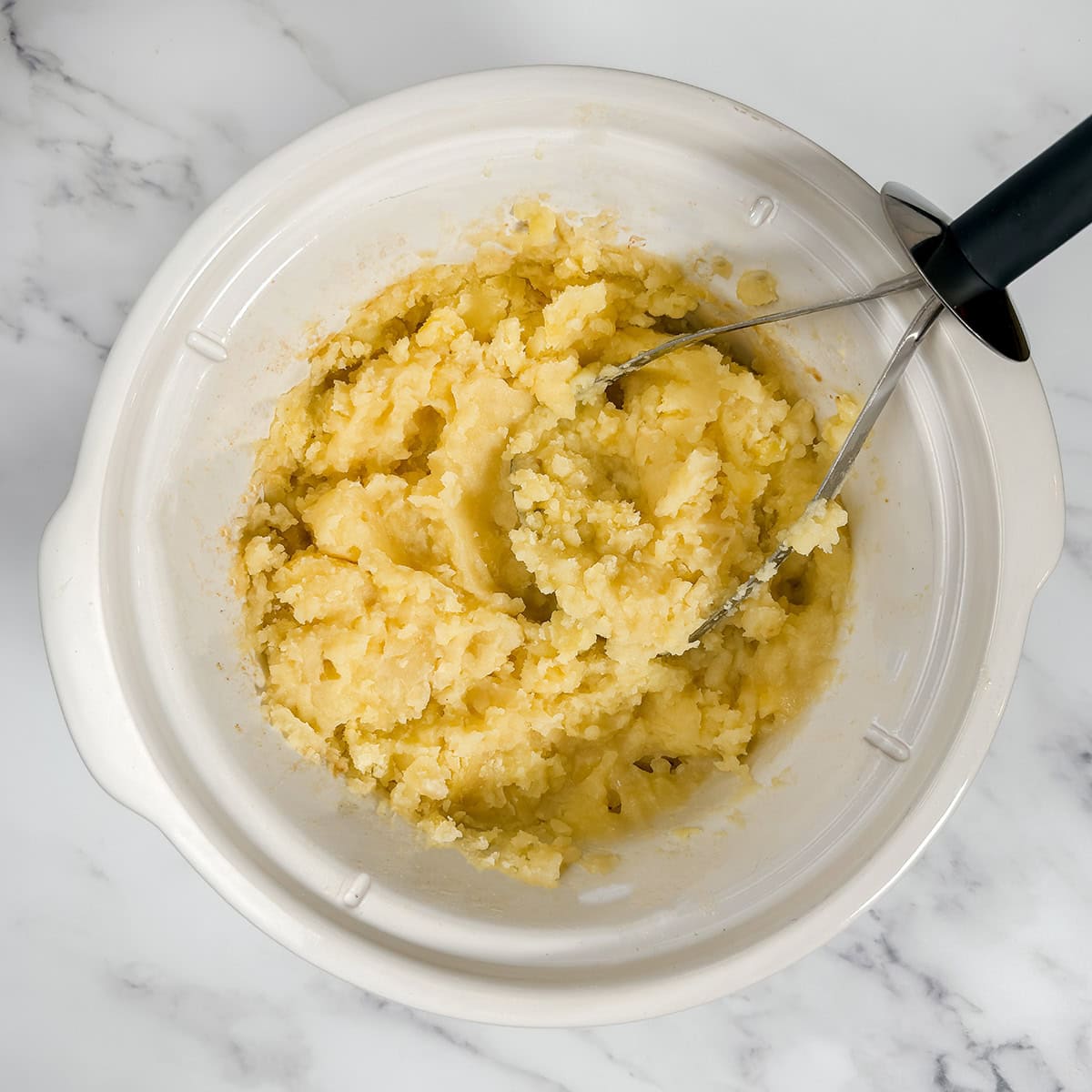 Mashing cooked potatoes in a crockpot with a potato masher.