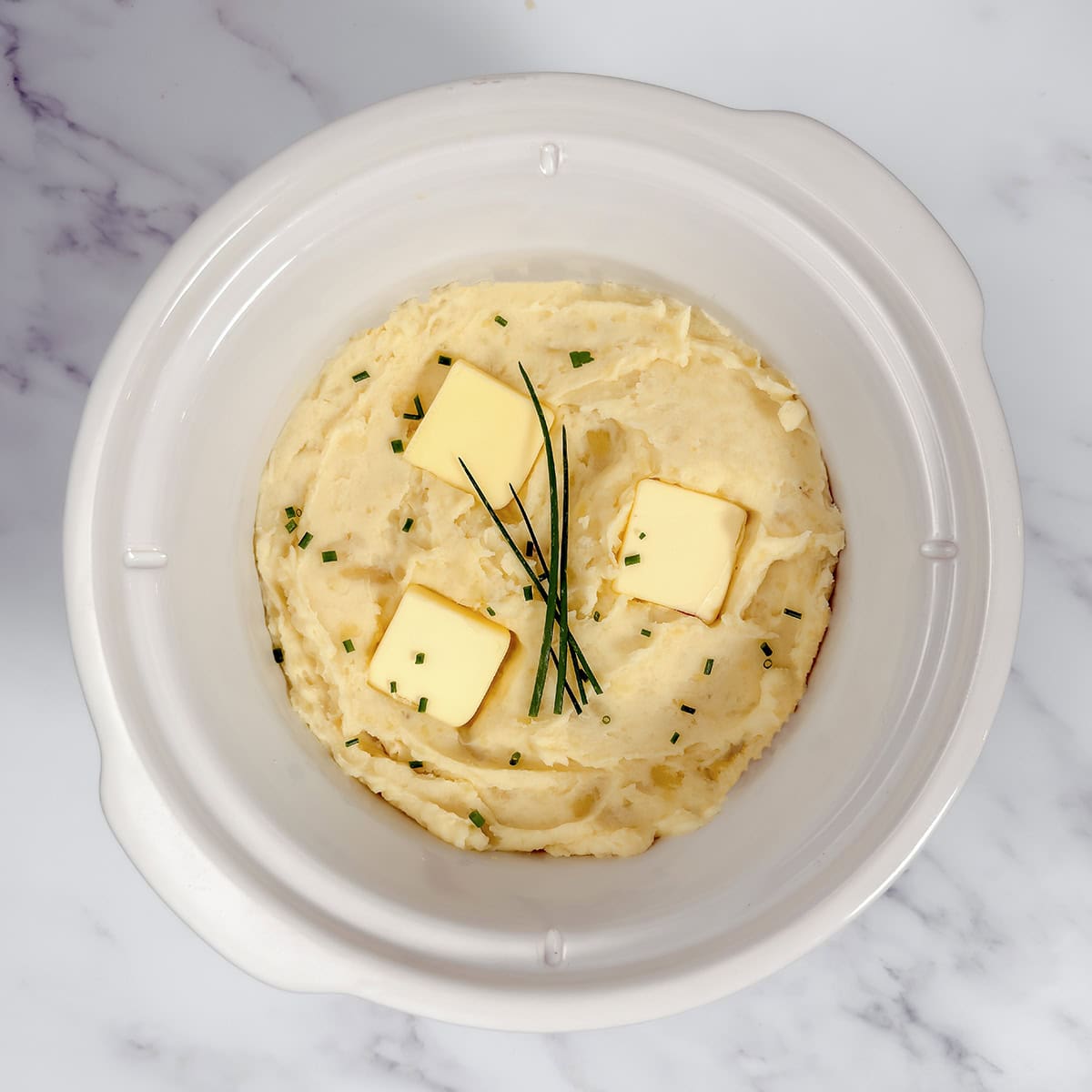 Freshly whipped mashed potatoes in a crockpot insert topped with butter pats and fresh chives.