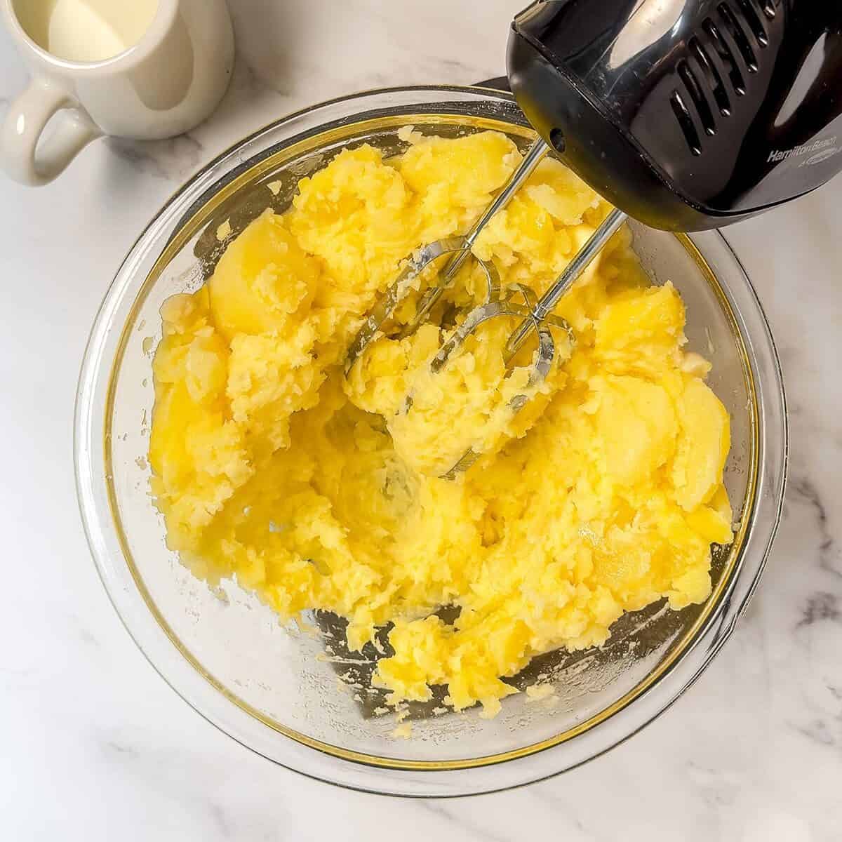 Cooked potatoes and garlic in a large glass bowl being mashed with a hand mixer.