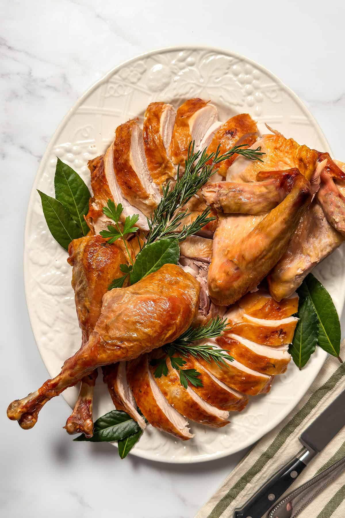 Sliced roasted turkey arranged on a platter with drumsticks, wings, and fresh herbs for serving.