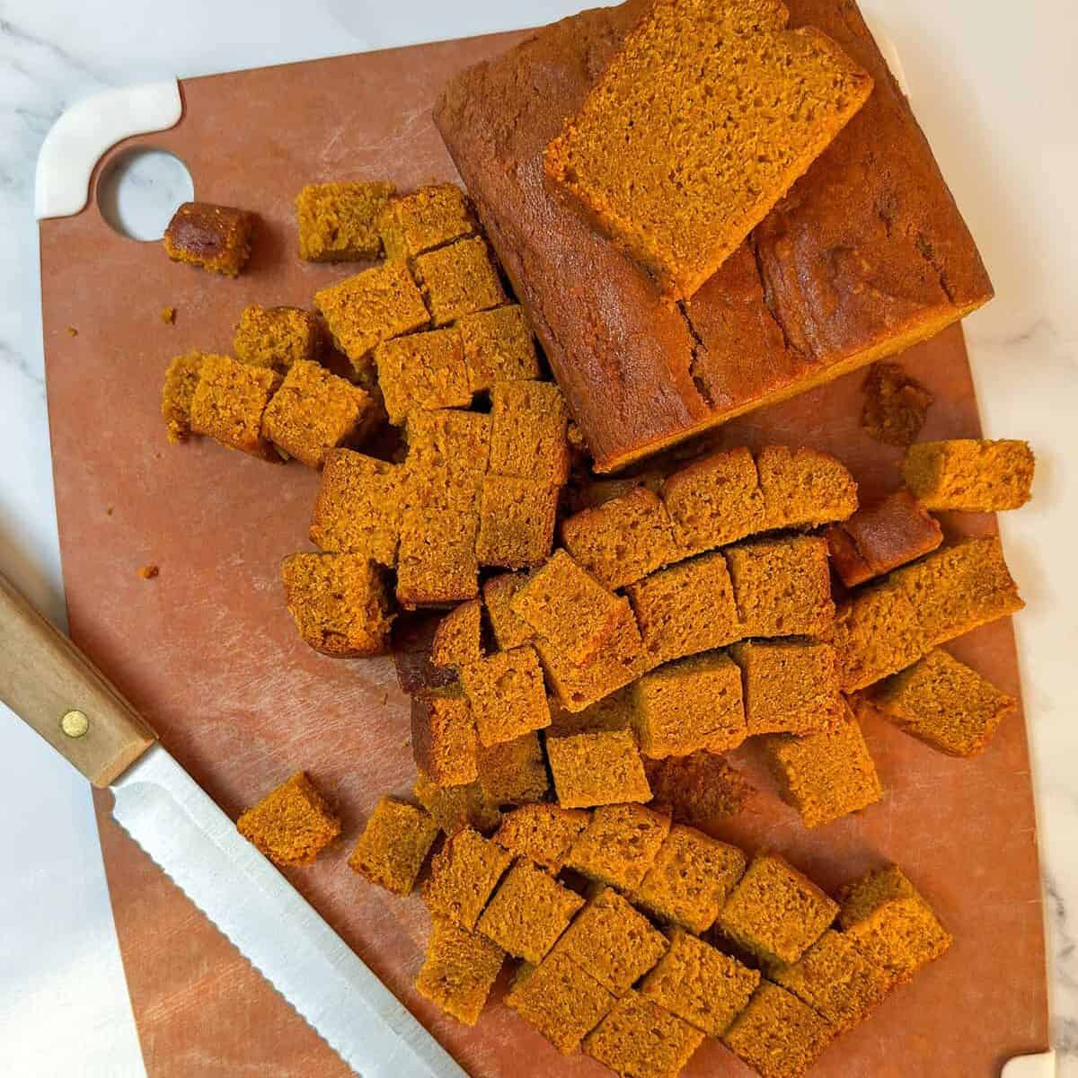 Cubes of pumpkin bread on a cutting board with a serrated knife.