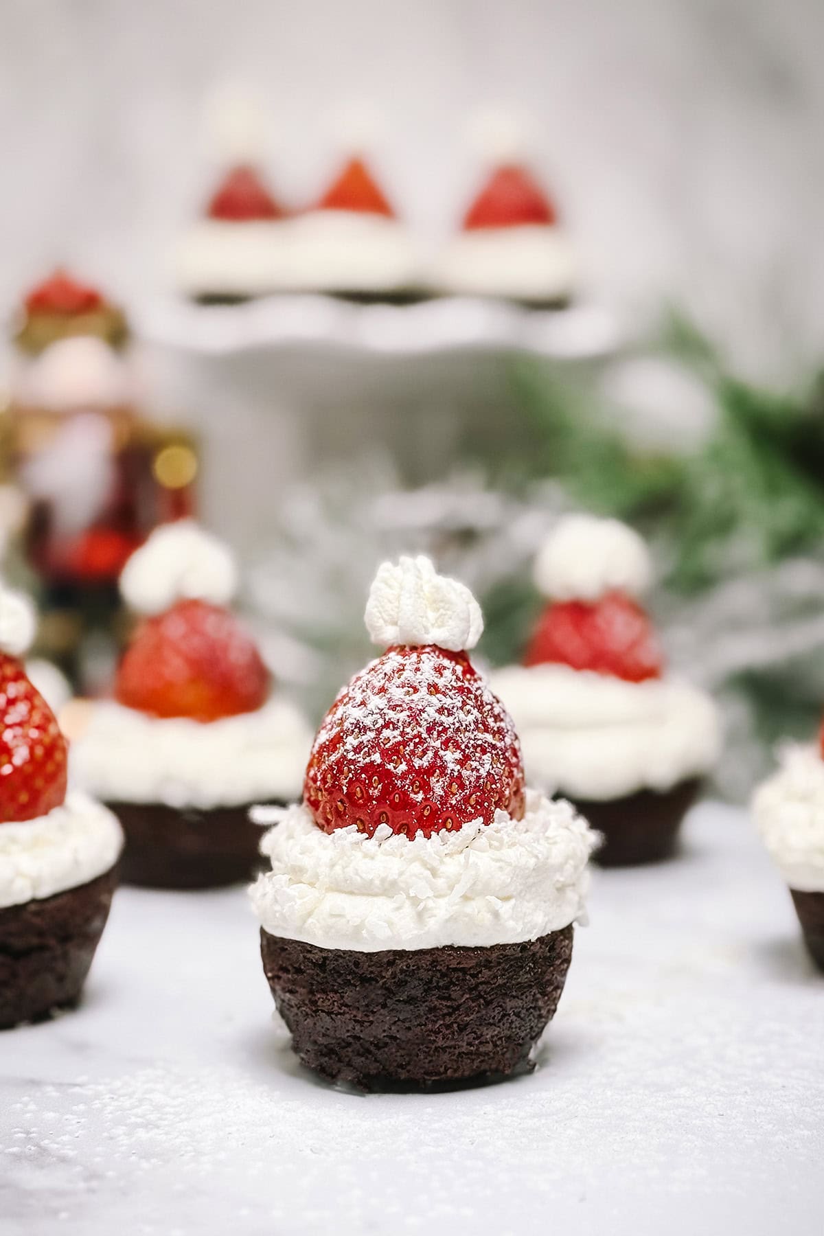 Santa Hat Brownie Bite topped with whipped cream and a strawberry dusted with powdered sugar, with festive holiday decorations in the background.