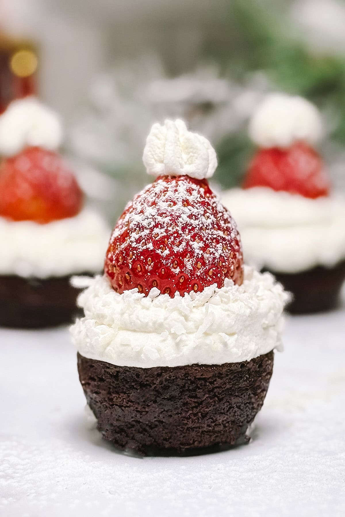 Closeup brownie bite topped with a powdered sugar dusted strawberry with more in the background. 