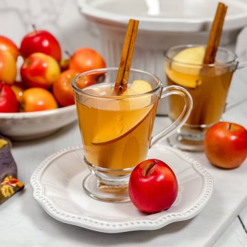 Square image of apple cider in glass mug garnished with cinnamon stick and apple slice a crockpot in the background.