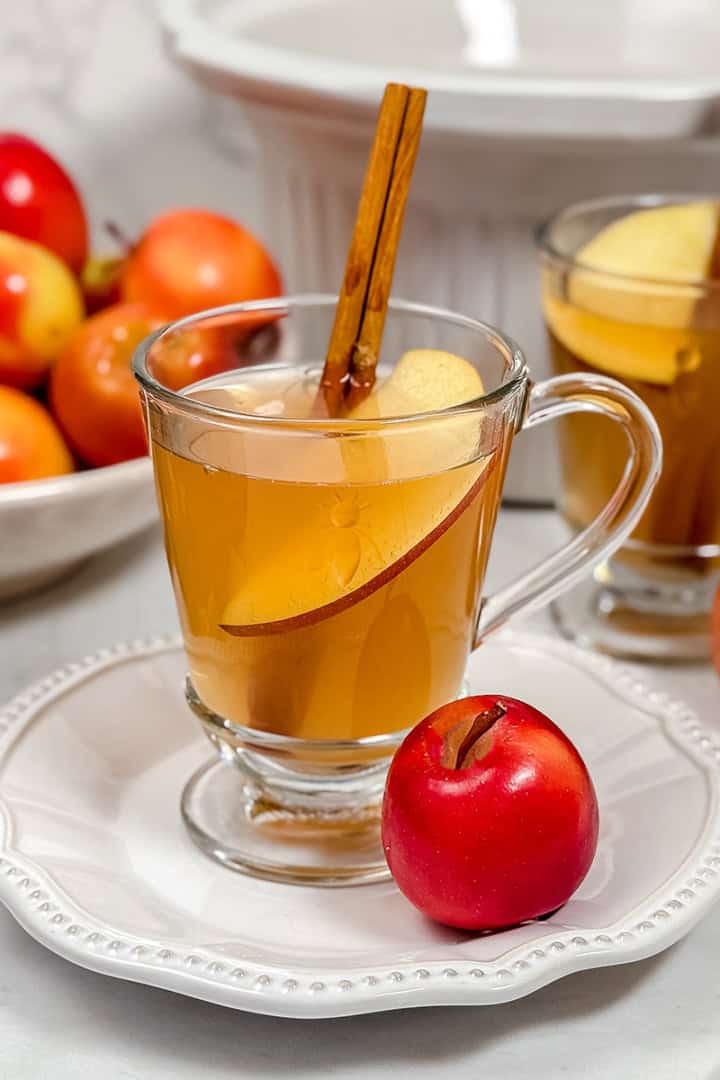 Square image of apple cider in glass mug garnished with cinnamon stick and apple slice a crockpot in the background.