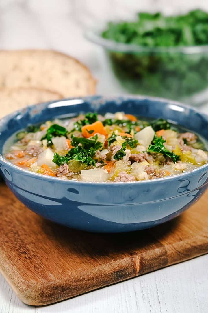 Square image of lamb and barley soup in a blue bowl with kale and sourdough bread in the background.