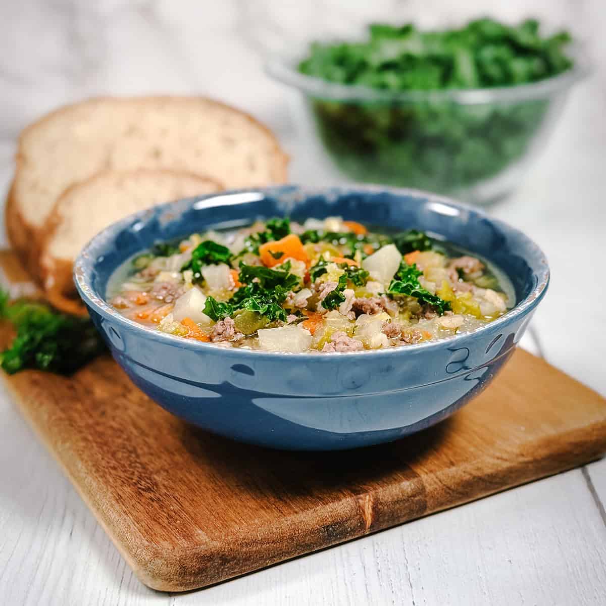 Square image of lamb and barley soup in a blue bowl with kale and sourdough bread in the background.