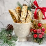 White cup holding Cranberry Pistachio Biscotti drizzled with white icing, surrounded by pine branches, pine cones, and festive holiday decorations.