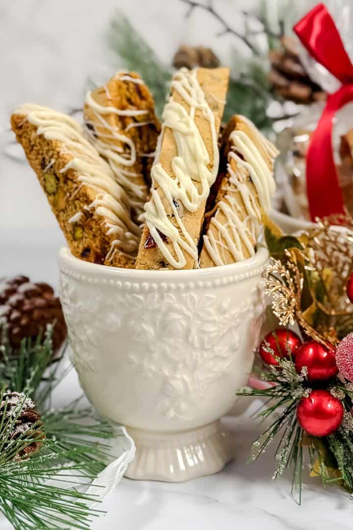 White cup holding Cranberry Pistachio Biscotti drizzled with white icing, surrounded by pine branches, pine cones, and festive holiday decorations.