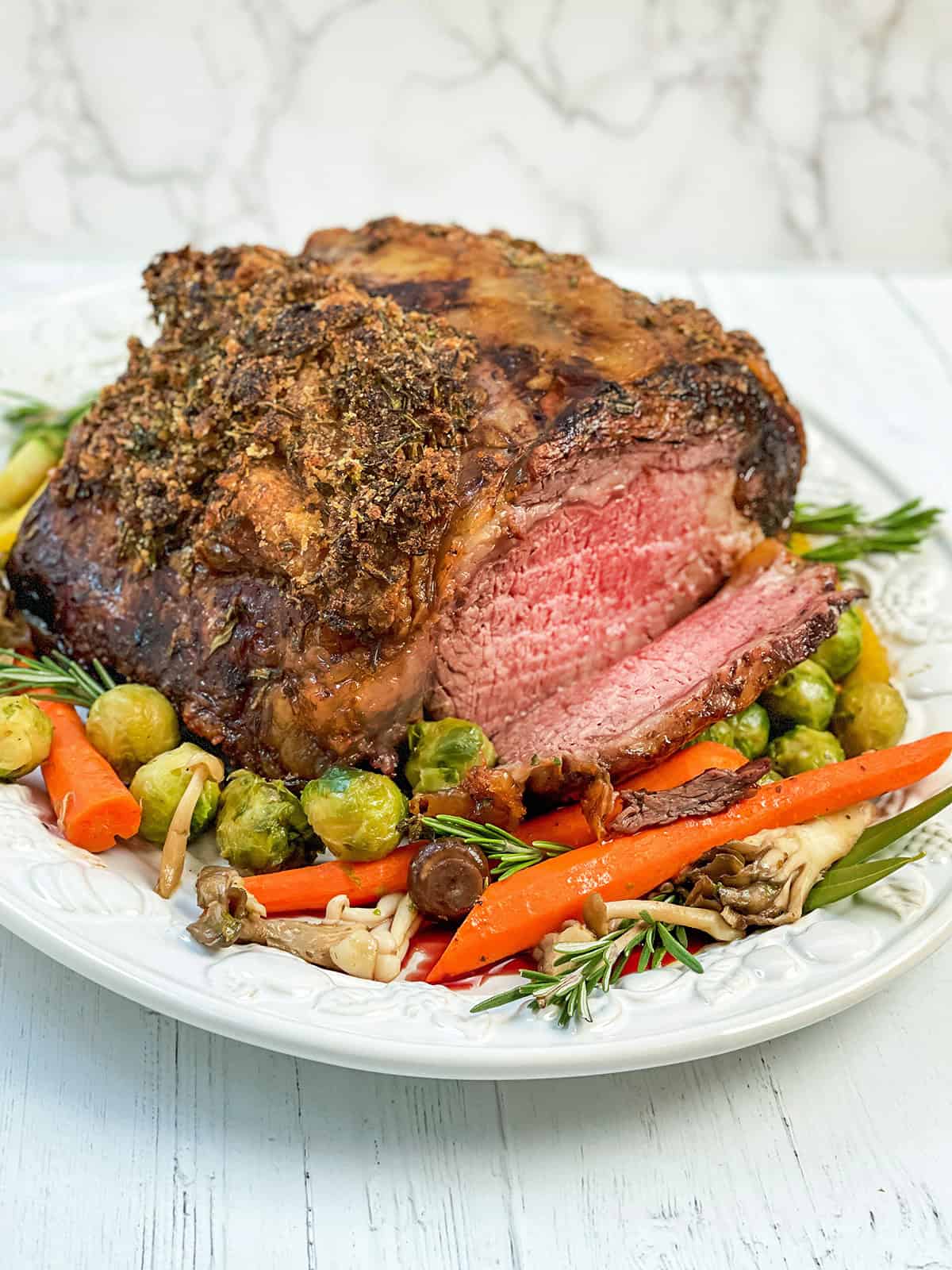 Prime Rib roast with herb crust sliced and resting on a serving platter with roasted vegetables for serving.