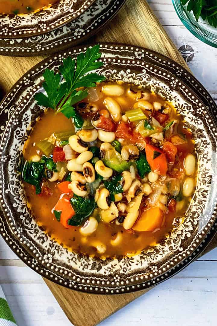 Bowl of New Year's Day Black Eyed Peas Soup in a patterned bowl and plate with freshly ground pepper and greens on the side.