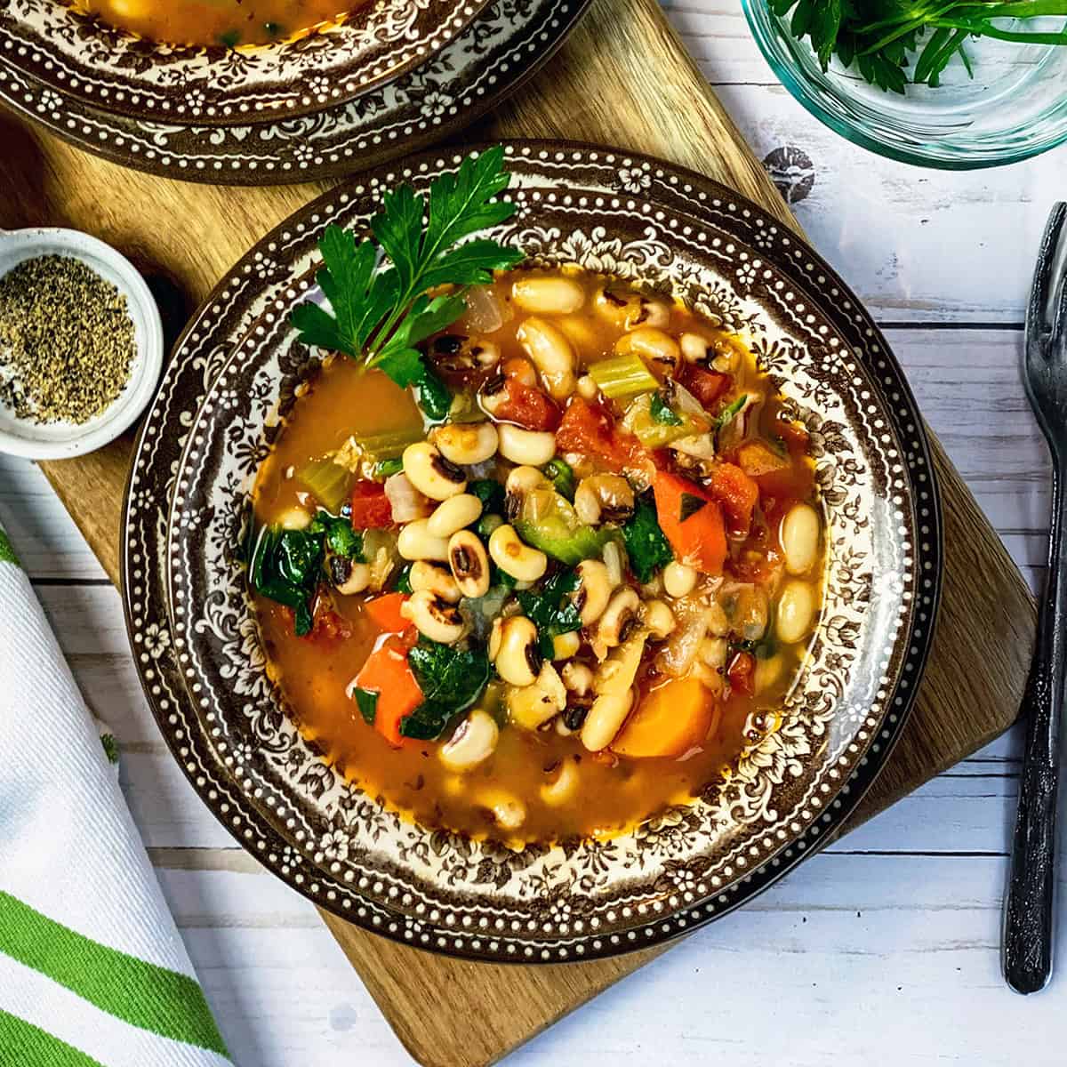 Bowl of New Year's Day Black Eyed Peas Soup in a patterned bowl and plate with freshly ground pepper and greens on the side.