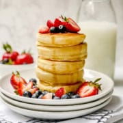 Tall stack of fluffy pancakes with cut fresh fruit on top and maple syrup drizzling down the sides.