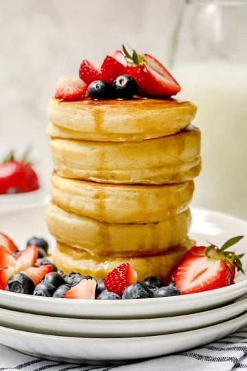 Tall stack of fluffy pancakes with cut fresh fruit on top and maple syrup drizzling down the sides.