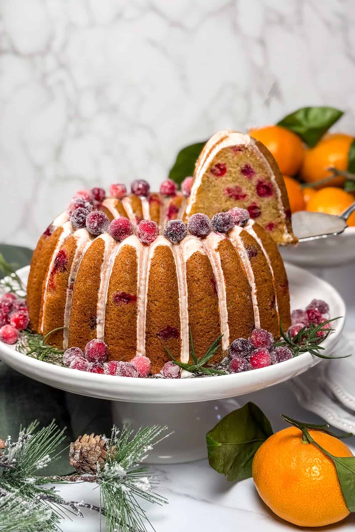 Cranberry orange Bundt cake with orange glaze and sugared cranberries, shown with a slice lifted to reveal the tender cranberry-studded crumb.