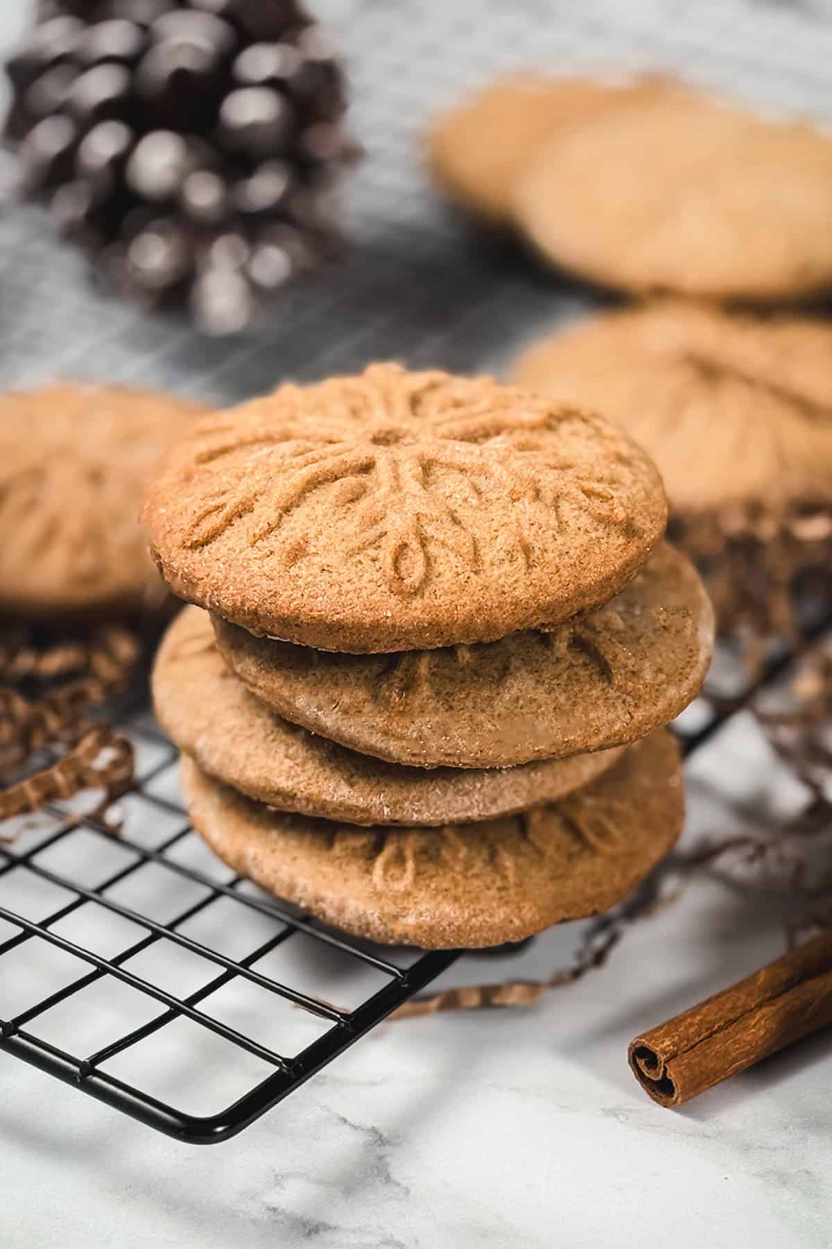 Stack of homemade speculoos cookies with embossed snowflake design, cooling on a rack with holiday d&eacute;cor in the background.