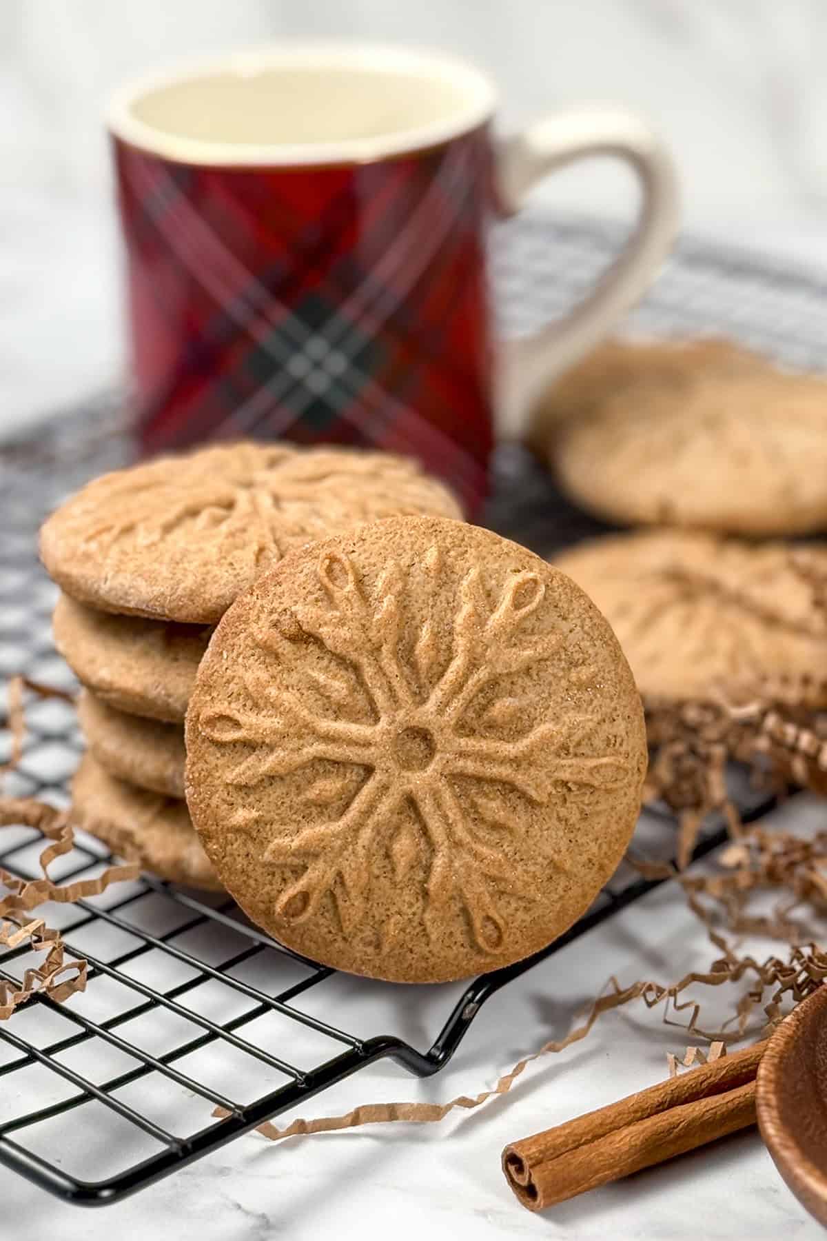 Speculoos cookies with snowflake stamp on a cooling rack next to a red plaid mug, perfect for St. Nicholas Day or holiday baking.