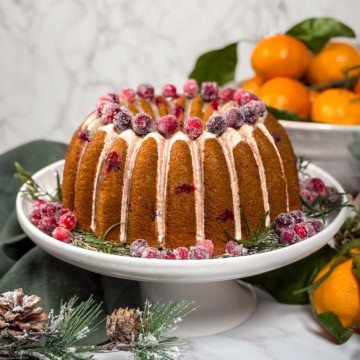 Cranberry orange Bundt cake drizzled with orange glaze and topped with sugared cranberries, displayed on a white cake stand with fresh oranges in the background.