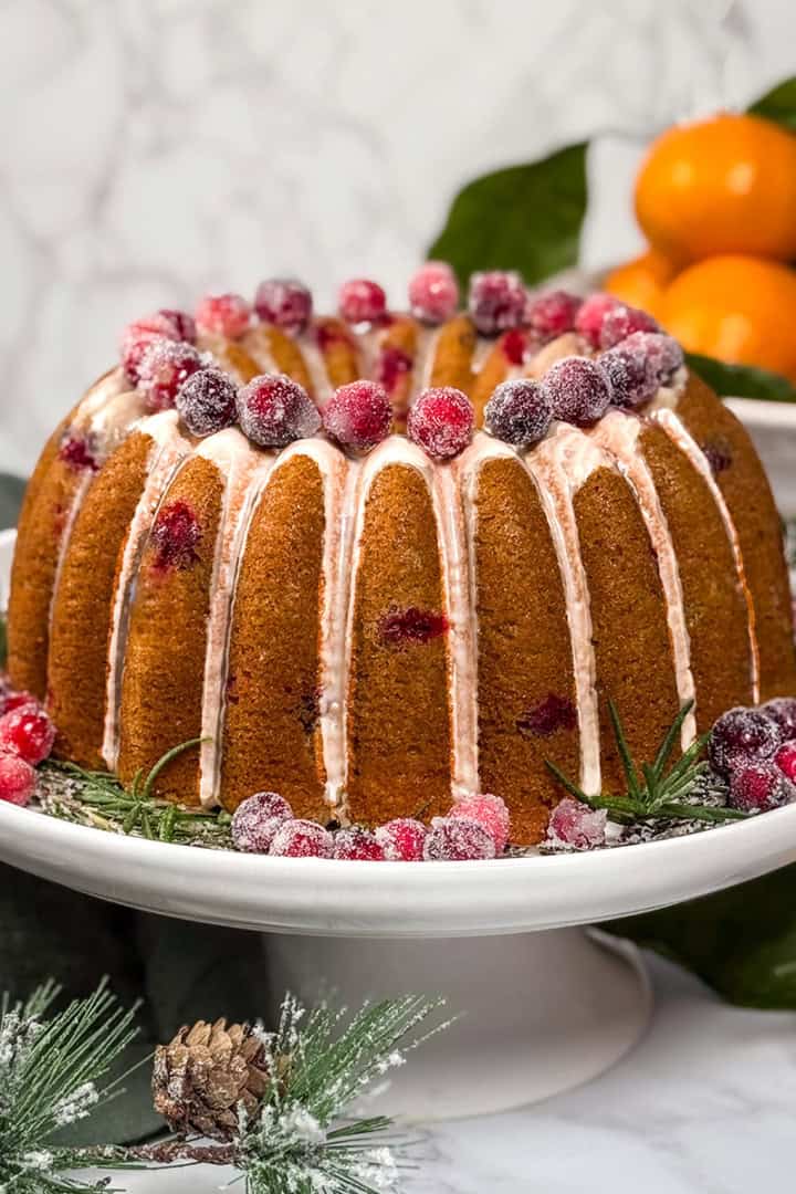 Cranberry orange Bundt cake drizzled with orange glaze and topped with sugared cranberries, displayed on a white cake stand with fresh oranges in the background.