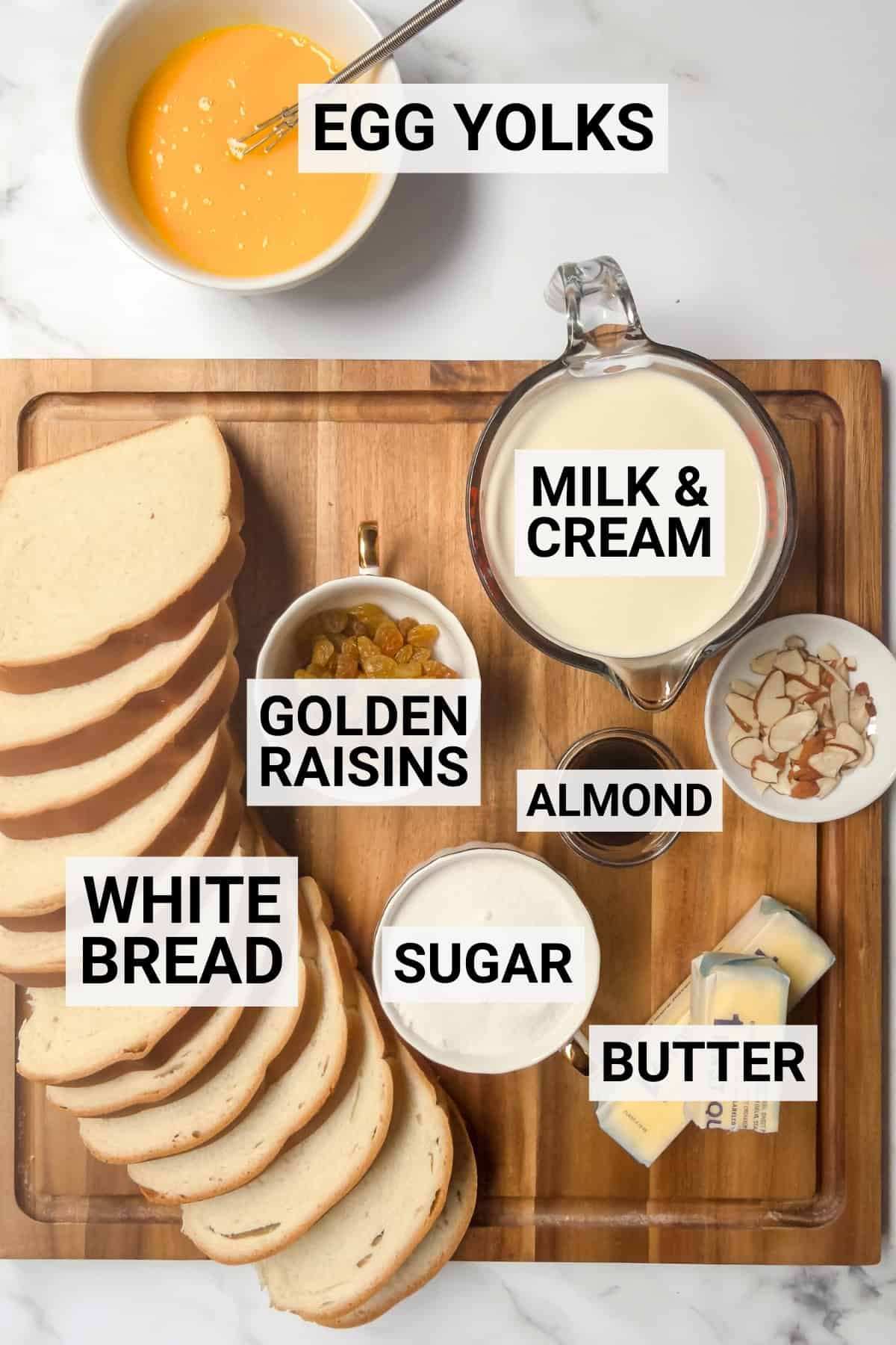 Ingredients for bread and butter pudding arranged on a wooden board, including sliced white bread, egg yolks, milk and cream, golden raisins, sugar, butter, and almonds.