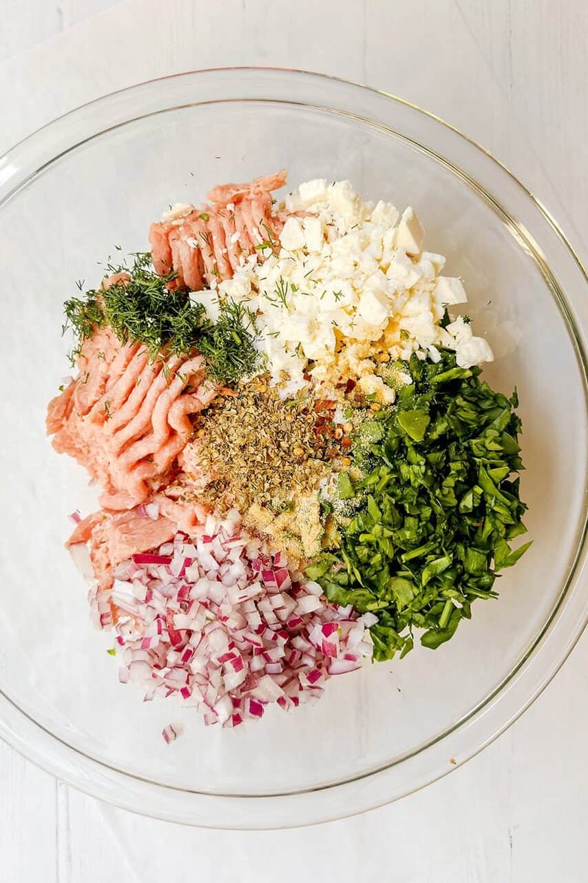 Greek turkey burger ingredients in a glass bowl with ground turkey, feta, herbs, spinach, and red onion.