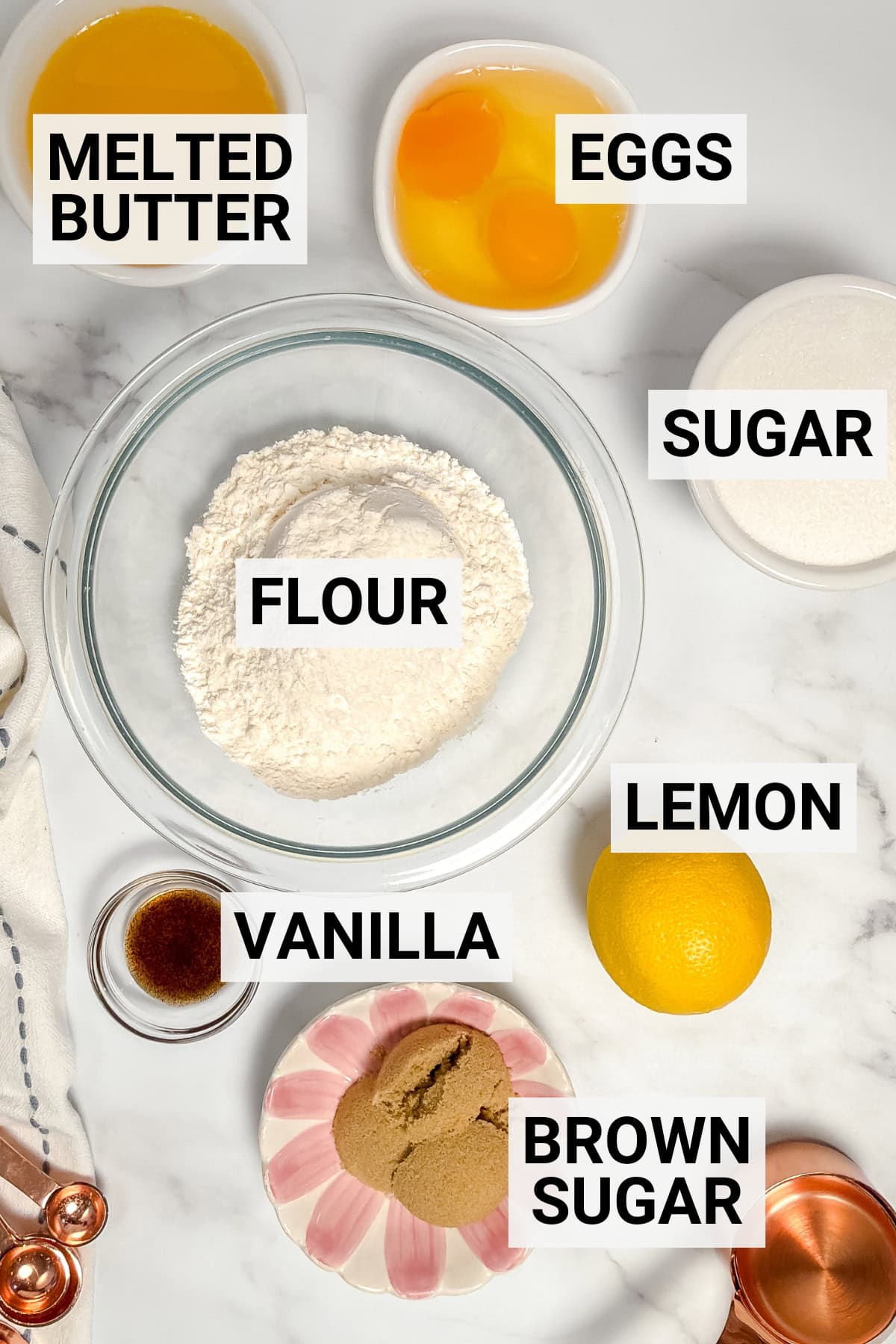 Top-down view of ingredients for a lemon madeleines recipe-melted butter, eggs, sugar, flour, lemon, vanilla, and brown sugar-arranged on a white surface and clearly labeled.