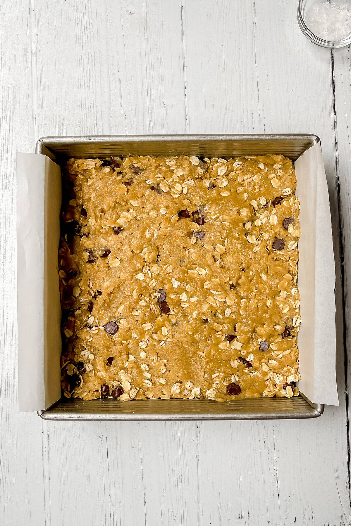 Pressing oatmeal chocolate chip cookie bar dough evenly into a parchment-lined pan.
