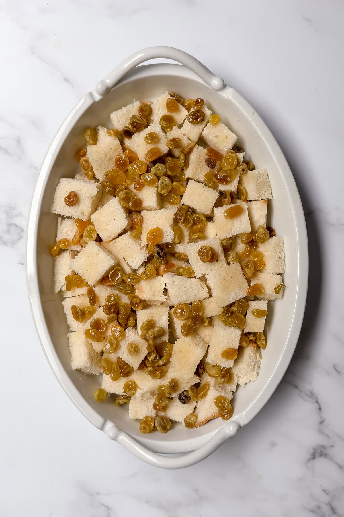 Golden raisins scattered over cubed bread in a baking dish for bread and butter pudding.
