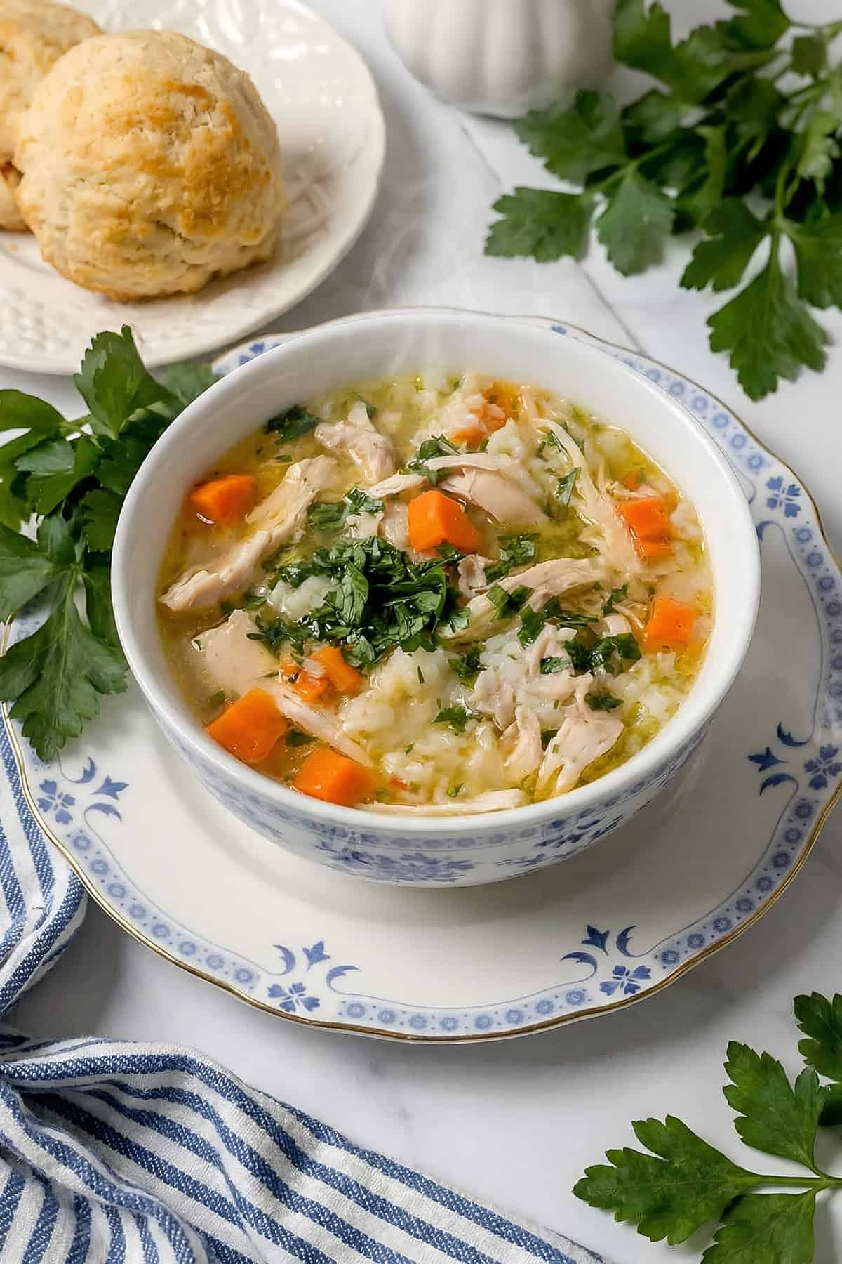 White bowl of chicken and rice soup topped with chopped parsley in a white bowl with homemade biscuits nearby.