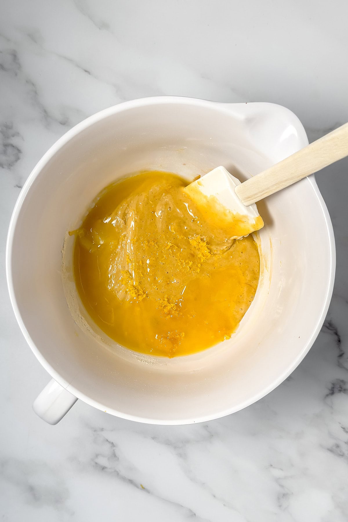 A white mixing bowl with Madeleines batter with melted butter around the edges and fresh lemon zest ready to be folded in.