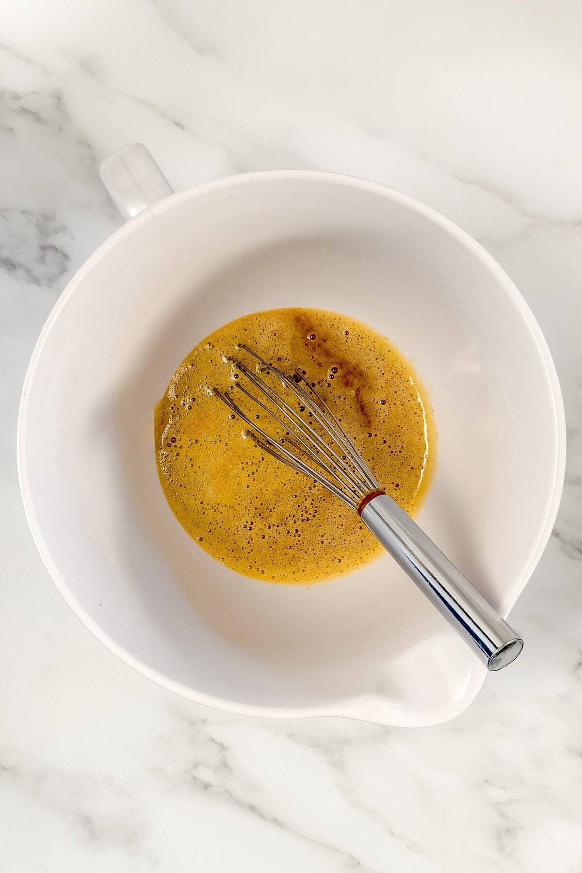 A metal whisk rests in a white mixing bowl containing a beaten egg mixture, the first step in crafting a delightful Lemon Madeleines Recipe, atop a white marble surface.
