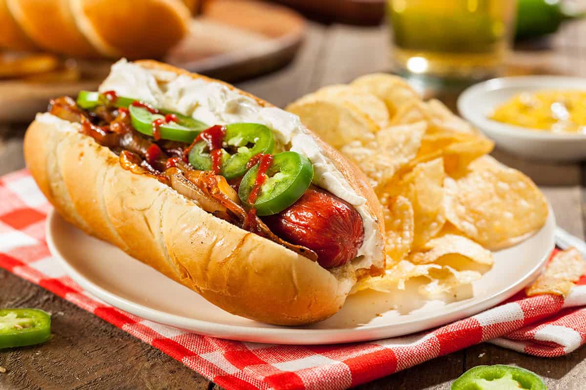 A Seattle-Style Hot Dog topped with cream cheese, grilled onions, jalape&ntilde;os, and ketchup on a bun, served with potato chips on a white plate and a red and white checkered napkin.