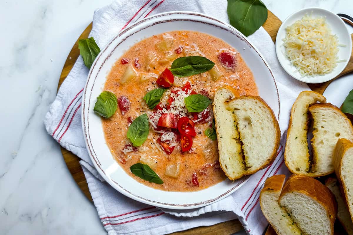 Bowl of tomato potato soup on a wooden board with bread and basil.